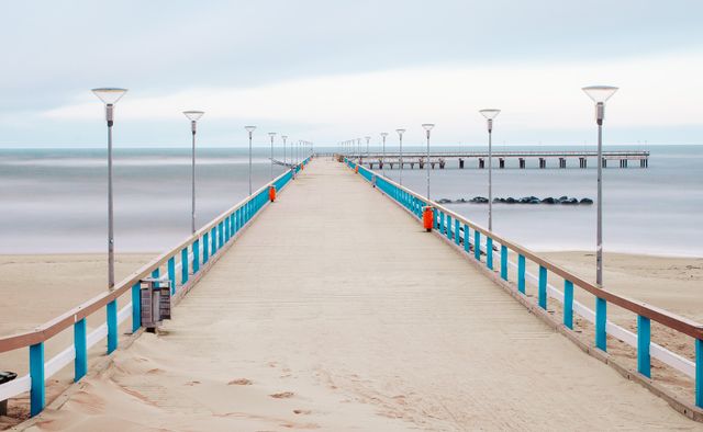 Pictured is an empty pier leading out to a calm ocean on a serene day. The scene captures the tranquility of the ocean beneath a clear horizon. This can be used for depicting themes of peace, solitude, or travel. Ideal for backgrounds in presentations, travel blogs, mental wellness content, or promotional material for beach destinations.