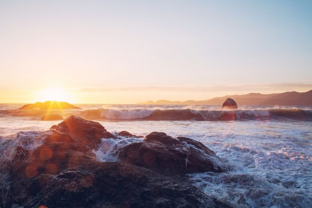 Beautiful sunset over a rocky seashore with waves crashing onto the rocks. The setting sun creates a warm glow on the horizon, casting colorful reflections on the ocean water. Perfect for use in travel brochures, nature calendars, desktop wallpapers, or inspirational quotes.