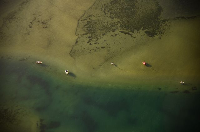 Aerial perspective of boats anchored in shallow, clear water along a coastline. Calm and serene ambiance depicting tranquil sea and shoreline with aquatic patterns. Ideal for use in travel and tourism promotions, nature documentaries, relaxation themes, and scenic coastal or nautical presentations.
