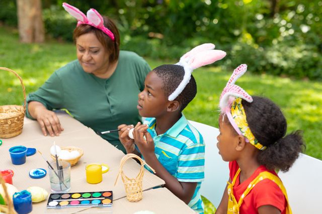 African American siblings and their grandmother wearing bunny ears are painting Easter eggs together outdoors. This image is perfect for illustrating family bonding, Easter celebrations, and creative activities. It can be used in articles, advertisements, or social media posts related to holiday traditions, family gatherings, and cultural celebrations.
