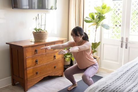 Young woman doing squat workout on yoga mat in bright plant-filled bedroom for fitness