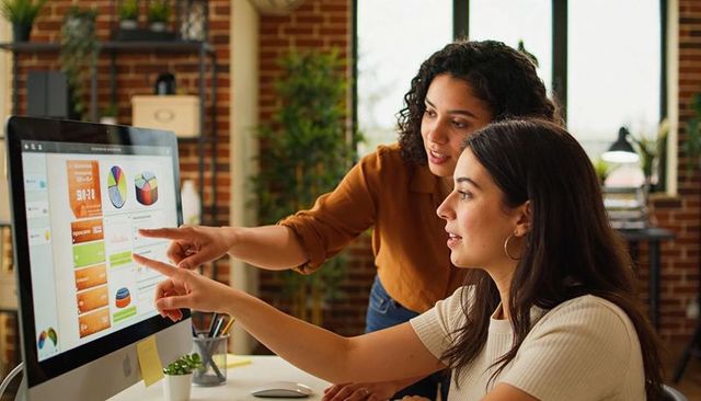 Two female colleagues collaborating on interactive data dashboard at modern brick-wall office desk
