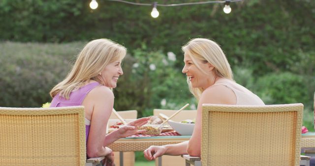 Two women engaging in a pleasant conversation while having dinner outdoors with string lights creating a cozy atmosphere. Ideal for themes around friendship, casual gatherings, outdoor dining, and leisurely summer evenings.