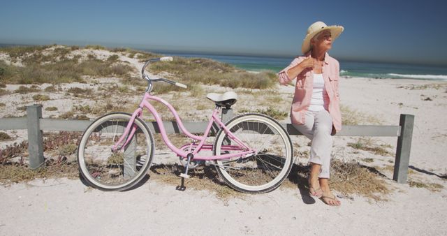 Elderly woman sits on a wood fence beside pink bicycle on the beach. Use to portray relaxation, summer vibes, seaside leisure or active lifestyle for seniors.