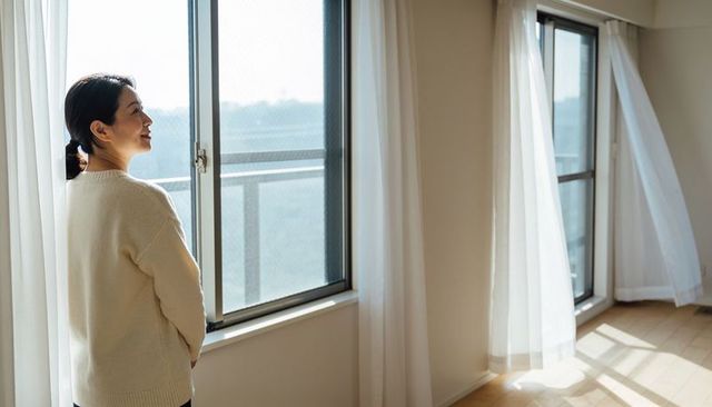 Asian woman gazing out sunlit balcony through sliding windows in minimalist apartment