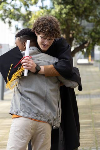 Diverse graduate hugging friend while holding diploma and mortarboard on campus