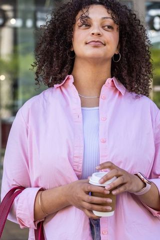 Confident african american woman holding to-go coffee wearing pink shirt in urban daylight