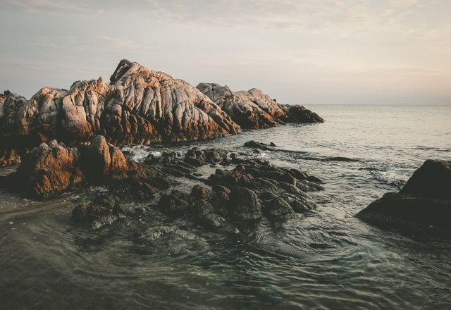 Picture showcases rocky sea coast during sunset with gentle waves lapping the shoreline. Useful for coastal and nature-themed projects, travel blogs, and tranquil environment backgrounds.