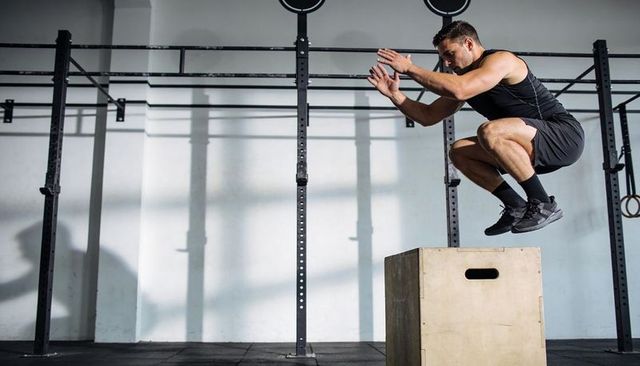 Explosive box jumping athlete tucking knees over wooden plyo box in industrial gym