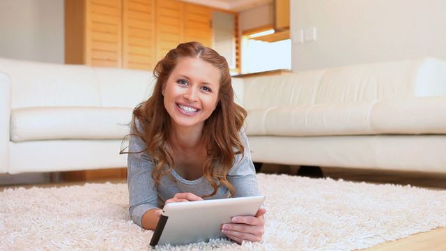 Young woman lying on a comfortable rug, engaging with a tablet. Ideal for conveying relaxation, modern technology, and home lifestyle themes. Suitable for promotions of technology, apps, and home furniture.