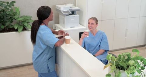 Diverse nurses chatting over coffee at clinic reception desk, healthcare teamwork