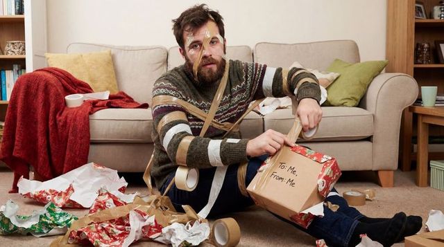 Bearded man unwrapping self gift amid tape and torn wrapping paper on living room floor