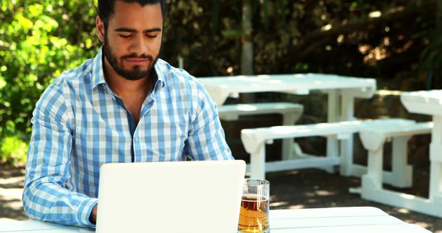 A young Middle Eastern man is focused on his laptop at an outdoor setting, with copy space. His casual attire and the presence of a beverage suggest a relaxed work environment or a leisure activity.