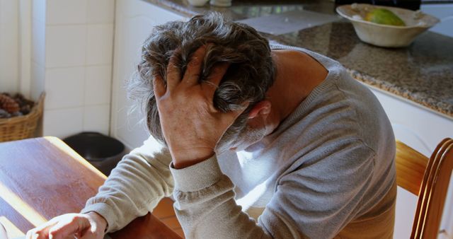 Mature man sitting at kitchen table with head in hands, expressing stress and frustration. Suitable for use in articles on mental health, aging, stress management, dealing with anxiety, emotional well-being, and life challenges faced by older adults.