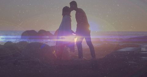 Sunset silhouette couple holding hands and kissing on rocky beach with lens flare