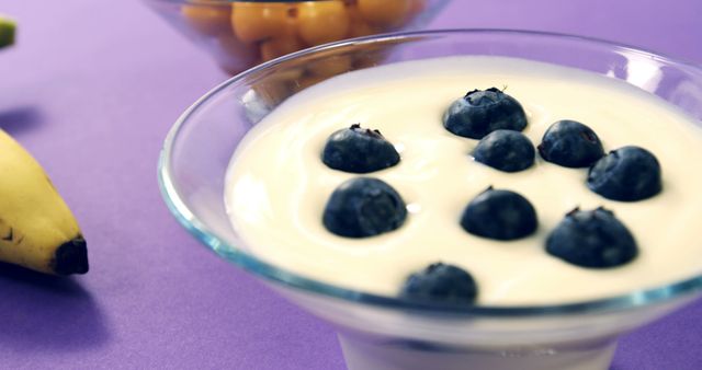 A bowl of creamy yogurt topped with fresh blueberries sits on a purple surface, accompanied by a banana and more fruit in the background. This setup suggests a focus on healthy eating or a nutritious breakfast option.