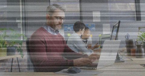Mature professional typing on laptop at shared wooden desk in modern open-plan office