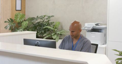 Clinic receptionist wearing scrubs and lanyard checking computer at modern healthcare desk