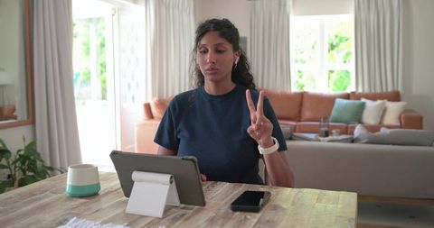African American woman doing remote video call on tablet, showing peace sign in cozy lounge
