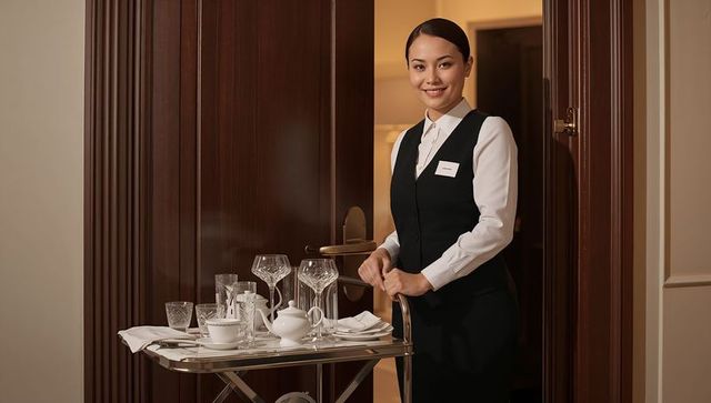 Hotel attendant in uniform standing with service cart and crystal glassware in room doorway