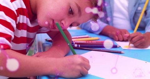 Leaning boy drawing at blue classroom table with green pencil, focused young artist