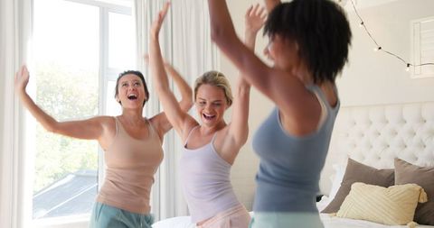 Diverse female friends dancing with joy in well-lit bedroom