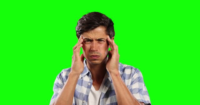 Young man in casual shirt holding head with both hands, showing signs of pain and stress. He seems to be having a headache or deep in thought, against a bright green screen background, making it perfect for chroma key editing. This can be used in various health, medical, or stress-related projects and articles highlighting mental health, modern life stress, or problem-solving.