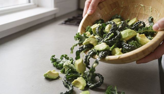 Pouring kale and avocado with creamy dressing from wooden bowl onto counter