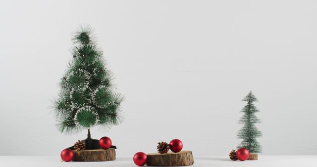 This image showcases two miniature Christmas trees placed on wooden slices, surrounded by red ornaments and pine cones, arranged against a plain white backdrop. It captures a minimalist holiday decor theme perfect for seasonal online content, advertising, social media posts, or blogging about decorating small spaces for Christmas or tackling DIY holiday crafts.