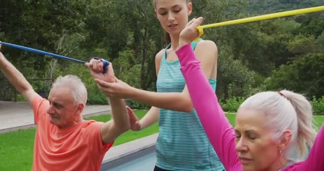 Senior couple is using resistance bands to exercise under the guidance of a young female personal trainer in a garden. Ideal for content related to fitness, elderly health, senior wellness programs, outdoor exercises, and personal training routines.