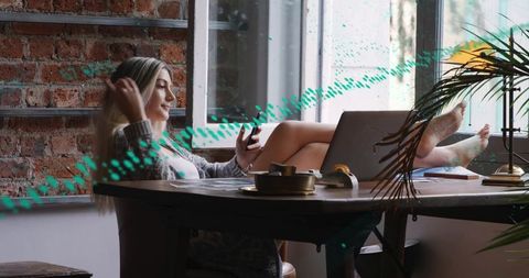 Reclining woman in loft workspace scrolling smartphone brushing hair with laptop in daylight