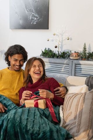 Couple laughing while opening gift on cozy holiday sofa with warm festive decor
