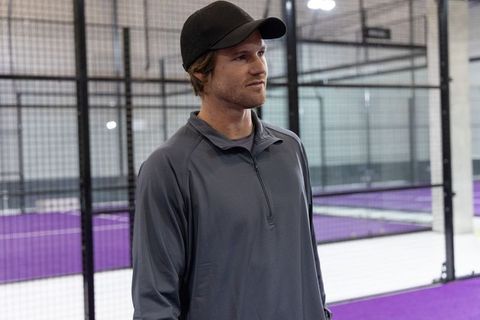Male athlete standing on indoor padel court wearing cap and gray training pullover