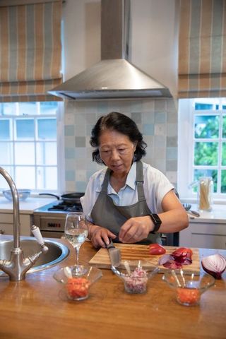 Senior chinese woman chopping vegetables on kitchen island wearing grey apron making meal