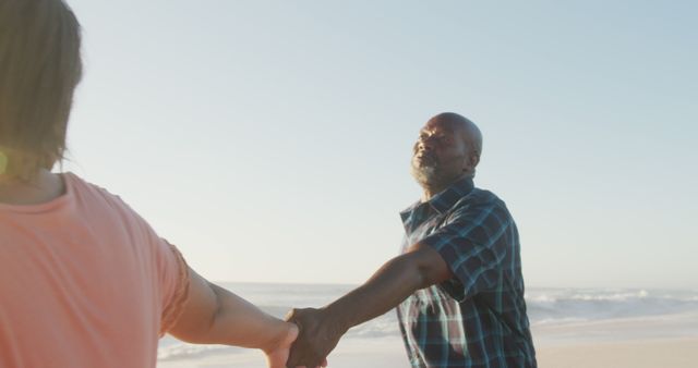 Smiling senior african american couple holding hands and walking on sunny beach. healthy, active retirement beach holiday.