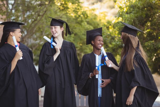 Group of diverse students celebrating graduation outdoors, holding diplomas and wearing caps and gowns. Ideal for use in educational materials, school brochures, graduation announcements, and articles about academic achievement and student life.