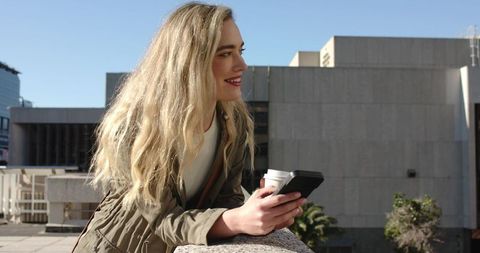 Young woman leaning on rooftop railing holding smartphone and coffee, smiling in city