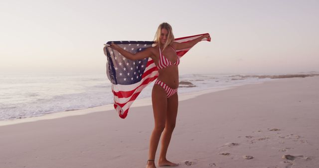 Young woman standing on beach holding American flag in red and white striped bikini. Creates a patriotic, summery feel perfect for content related to summer holidays, beach trips, or American pride. Ideal for promoting travel destinations, patriotic events, beachwear fashion, and summer campaigns.