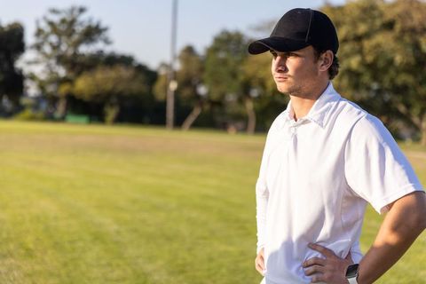 Young man standing hands on hips on sports field wearing cap and polo during golden hour