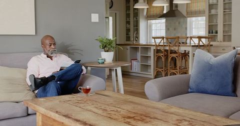 African man relaxing on sofa reading book with tea in cozy living space