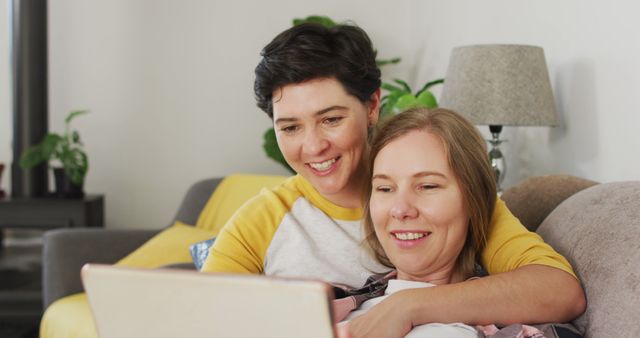 Happy LGBTQ couple relaxing at home, enjoying time together with a laptop. One person has short dark hair, wearing a casual shirt, while the other has short light brown hair, resting on the sofa. Cozy and intimate setting invokes feelings of love, happiness, and togetherness in a casual environment. Useful for themes related to LGBTQ relationships, technology in daily life, and domestic bliss.