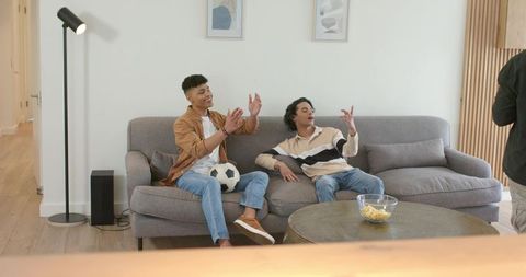 Diverse friends cheering on sofa while watching soccer game at home, holding soccer ball