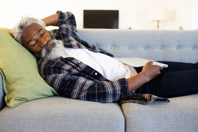 This image depicts an African American man resting on a sofa with a laptop in his hand, suggesting a relaxed and comfortable home environment. Ideal for use in articles or advertisements related to home lifestyle, relaxation, remote work, or quarantine life.