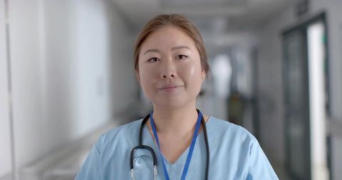 Standing nurse in light-blue scrubs wearing stethoscope and lanyard in hospital corridor
