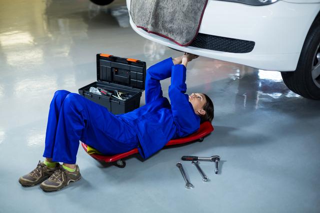Woman in blue overalls lying on a creeper under a white car, fixing components with various tools around her. Perfect for illustrating auto repair services, women in technical professions, and car maintenance advertisements.