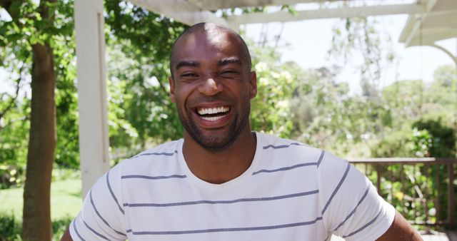 This image shows a cheerful African American man smiling broadly while outdoors in a sunny park. The man is wearing a casual white shirt with stripes, and background comprises greenery and trees, suggesting a pleasant, warm day. Suitable for use in promotional materials for products and services emphasizing joy, outdoor activities, health, happiness, and leisure. Could also be used in blog posts or articles focused on lifestyle, well-being, and diversity.