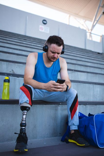 This image depicts a Caucasian male athlete with a prosthetic leg sitting on stadium stairs, using a smartphone. He is wearing sportswear and headphones, with a water bottle and sports bag nearby. Ideal for use in articles or advertisements promoting inclusivity in sports, athletic perseverance, or technology in fitness.