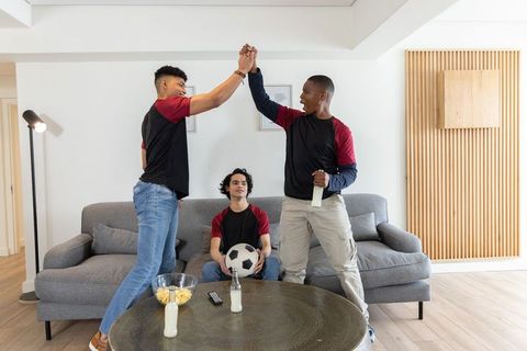 Friends celebrating soccer win in modern living room high-fiving on sofa
