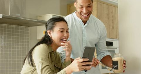 Mixed-race man and Asian woman laughing while checking smartphone in bright cozy kitchen