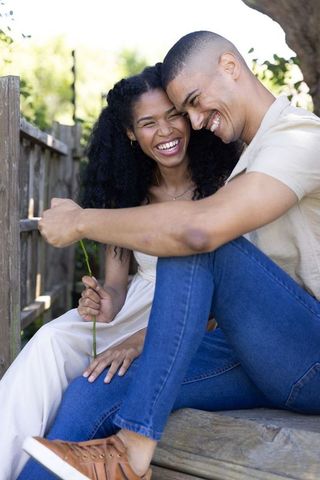Laughing couple leaning together on bench in backyard holding flower, candid affection